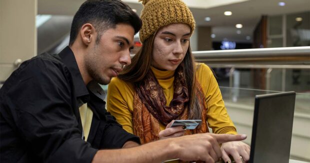 Young Latin American couple hold a credit card in their hand and use a laptop and cell phone to make Internet payments