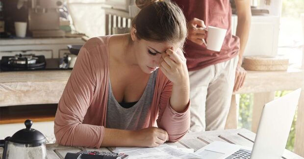 Stressed young female sitting at kitchen table with papers and laptop computer